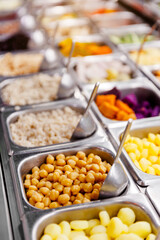 Close-up of a buffet display featuring chick peas, peanuts, and white beans in stainless steel trays, with vibrant vegetables in the background. A colorful and healthy food variety.
