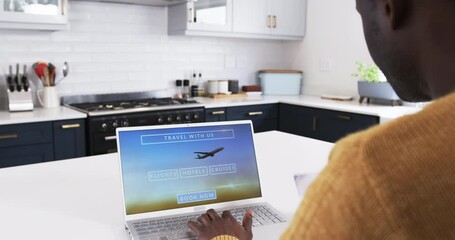 African American man booking holiday travel online using laptop in modern kitchen