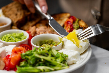 A close-up of a delicious brunch plate featuring fresh vegetables, guacamole, and poached eggs with runny yolks. A hand is seen cutting into the egg, creating an appetizing scene.