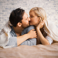 Fototapeta premium Portrait image - inlove kissing couple with closed eyes, covered with sheet-blanket over heads, lay on bed, against bricks wall background. Woman, man at home. Love, relationship, happy family concept