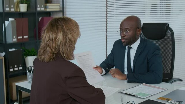 African American financial advisor in formal suit sitting at desk in office, giving contract to Caucasian female client for signing and explaining terms during consultation