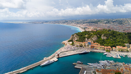 Obraz premium Aerial view of Nice, France, showcasing the coastline with turquoise waters, luxury yachts in the harbor, and a cityscape blending with lush greenery under a partly cloudy sky