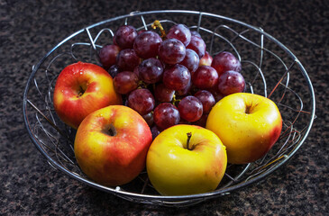 A metal fruit bowl contains four freshly washed apples and one branch of large red grapes, fruits of late-ripening varieties.