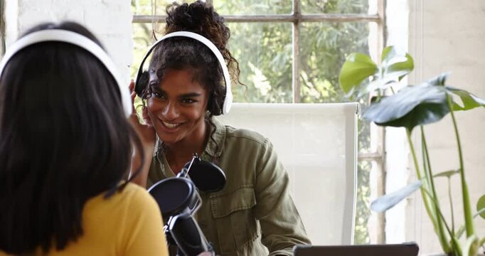 Two multiracial female colleagues recording podcast at home, wearing headphones and smiling warmly
