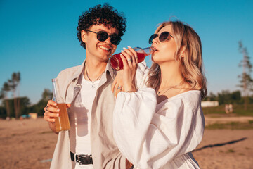 Young stylish friends posing at the seaside. Fashion man and cute female dressed in casual summer clothes. Smiling models having fun. Cheerful women and guy outdoors, Hold and drink lemonade