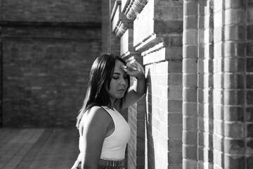 Obraz premium pretty young brunette woman rests her head on her arm on the wall and looks down. The woman is sad and depressed and feels pain and despair. She feels bad and negative. monochrome photo.