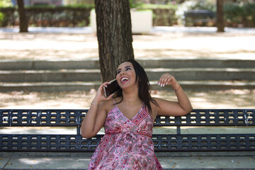 A brunette, young and beautiful woman is sitting on a park bench and talking on a mobile phone. The woman is happy and smiling with laughter and is having fun while listening. Concept of telephony.