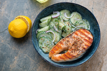 Blue bowl with cucumber salad and grilled salmon, horizontal shot on a grey and beige granite surface, elevated view