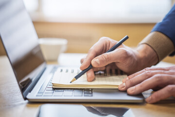 Sharp view of a man's hand transcribing in a notepad on a cutting-edge laptop, with a defocused backdrop