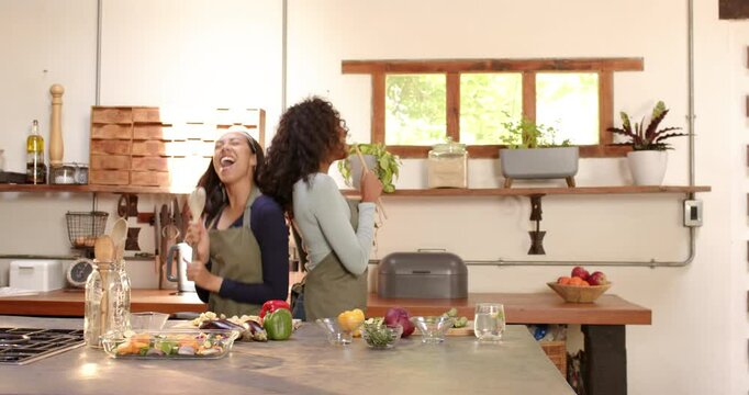 Two multiracial female friends joyfully singing and cooking together in cozy kitchen, at home