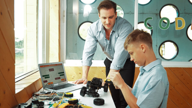 Caucasian teacher help student fixing car model and giving advise for programing system or coding prompt while highschool boy listen and use equipment to repair robot at STEM classroom. Edification.