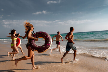 Group of happy young friends are having fun and runs to swim at the beach. Group of tourists with inflatable rings at sunset sea