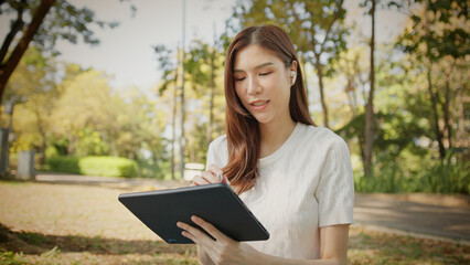 Obraz premium Asian Woman Working outdoors in the park. Professional on a Video Call, exploring Hybrid Work Opportunities in a Peaceful, Virtual Meeting and Online Learning. Hybrid work lifestyle concept by tablet