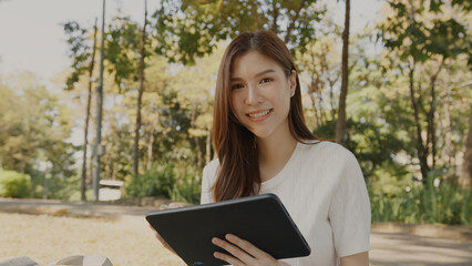 Fototapeta premium Asian Woman Working Remotely in a Park, Using Tablet for Hybrid Work Lifestyle, Tranquil Freelance, Joyful Digital Nomad in Nature, Relaxing and Working in Nature, Remote work using Tablet in greenery