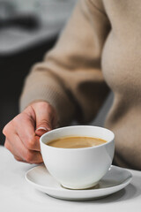 A close-up of a person's hand holding a white cup of coffee on a saucer. The background is softly blurred, creating an inviting and cozy atmosphere. Ideal for lifestyle or beverage themes.