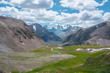 Fototapeta premium Mountain creek and lake in sunlit green alpine valley against few big snowy pointy peaks far away under clouds in blue sky. Awesome scenic aerial view to three large beautiful snow-capped peaked tops.