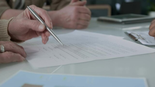 Close up view of hands of unrecognizable financial advisor pointing at key terms of contract and explaining details to senior woman before signing document at office meeting