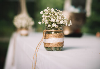 Wedding bouquet of white small flowers in a glass vase on a white fabric cotton tablecloth with green background.
