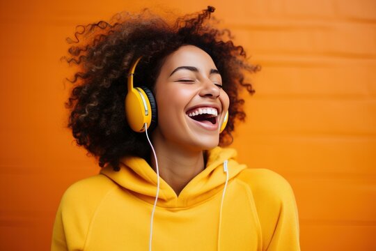 Carefree young woman with yellow headphones and hoodie enjoying music, laughing with closed eyes, on orange background