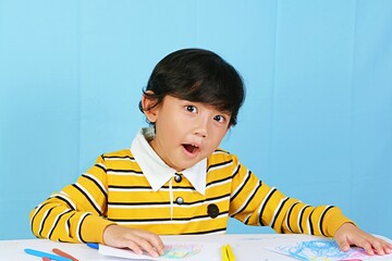 Fototapeta premium A young boy in a striped yellow shirt engaged in creative art activities with colorful crayons on a white table. Perfect for educational, childhood development, or creative learning themes.