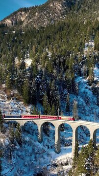 Aerial Vertical view of Landwasser Viaduct and Bernina express in Swiss Alps snow winter scenery. Drone shot red train passing through mountain in Filisur, Switzerland.
