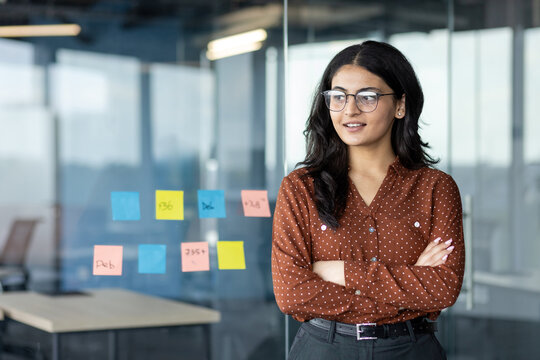 Young successful female office worker at workplace smiling with crossed arms looking towards the window. Businesswoman is satisfied working inside the office. - Powered by Adobe
