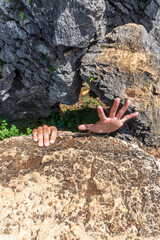 Two hands reach up from behind a rock formation, with an open palm, showing vulnerability and struggle.