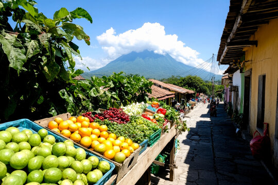 A vibrant market scene features fresh produce in baskets under a clear blue sky, with a majestic mountain in the background.