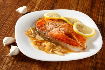 Grilled salmon steak with lemon slices and garlic on a white plate, set on a rustic wooden table.