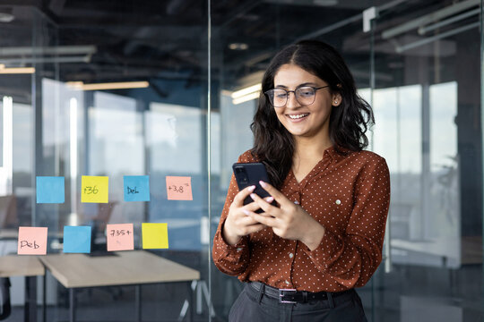 Young businesswoman successful office worker standing with phone in hands, typing message smiling happily. Woman in casual clothes standing by window.