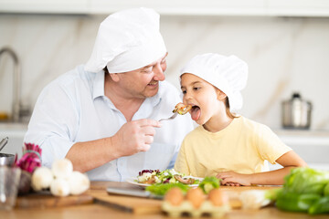 Father in a chef hat offers his little daughter to try the prepared salad in kitchen