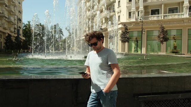 man with curly hair in a striped T-shirt and sunglasses with a phone in his hands near a fountain, Tashkent, Uzbekistan