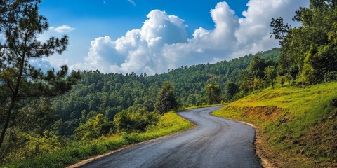 Paved road winding uphill through a serene forest, featuring a clear sky where clouds are distinctly visible. This image captures the beauty of a paved road in nature.