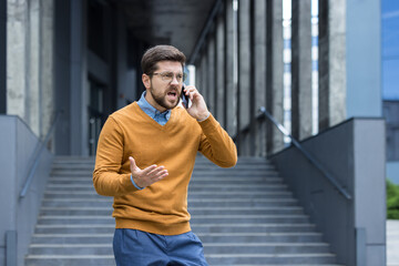 An angry young man is standing outside an office building and is talking on the phone while...