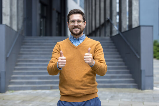 Portrait of a young successful man standing outside an office building smiling and showing the super gesture with his fingers to the camera