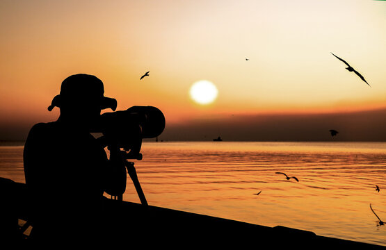 Silhouette of a photographer using  camera with large telephoto lens for shooting seagulls in the evening on sunset background. Vacation at Bangpu recreation center, Thailand the lifestyle of  hobbies