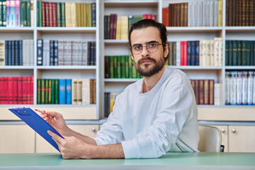 Portrait of young male teacher, psychologist with clipboard sitting at desk in library