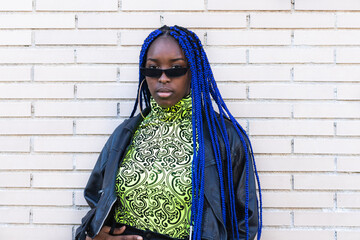 Confident African American female with blue braids wearing trendy outfit and sunglasses standing on...