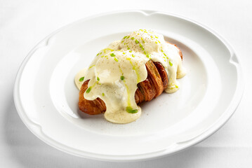 A delicious croissant topped with creamy hollandaise sauce and garnished with green onions on a white plate. Perfect for a gourmet breakfast or brunch setting.