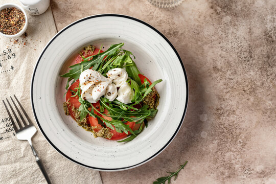 Caprese italian salad with fresh mozzarella, sliced tomatoes, aragula and pesto sauce served in white plate on beige background top view, text space