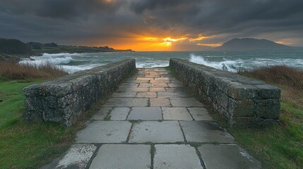 Fototapeta premium A serene pathway leads to a dramatic sunset over turbulent waves and distant mountains.