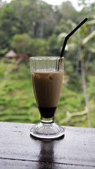Iced latte in a tall glass with a black straw on a wooden table with blurred tropical greenery and rice terrace view in Bali