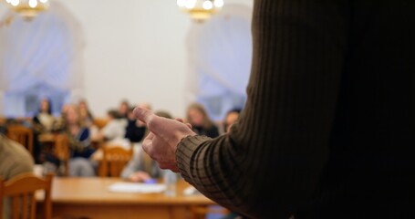 Man wearing brown turtleneck faces attentive crowd, delivering speech with purposeful intensity, emphasizing speech value in knowledge exchange, urging listeners to heed speech insights