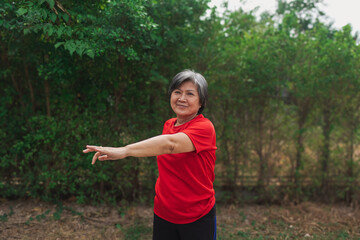 Happy asian old woman stretching arms and body at park in morning after exercise, wear red t-shirt, healthy routine.