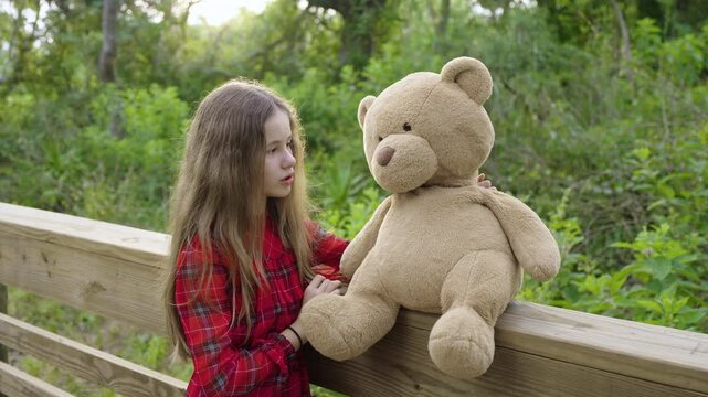Pretty preteen girl playfully taking to her teddy bear friend outdoors on wooden boardwalk in Florida jungle. Concept of friendship values