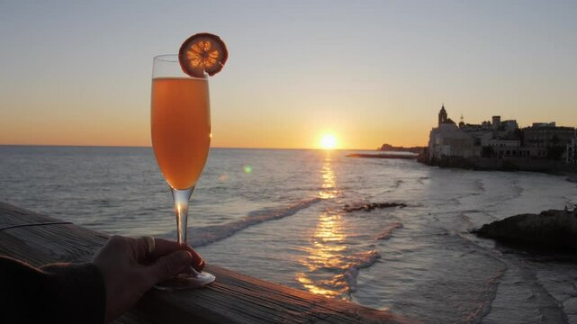 Glass of orange Bellini cocktail in woman hand, with sea and waves in background during golden hour. Romantic sunset date at cafe in historic European town Sitges.