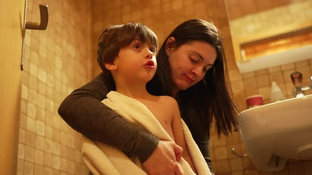 Mother kissing son on the cheek and drying body wrapped in towel after bath time. Authentic parent and child loving affection and caring relationship