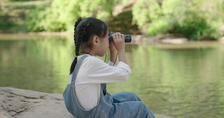 Asian girl sitting by a calm forest river, using binoculars for birdwatching, symbolizing curiosity, love for nature, and peaceful exploration in a serene environment with family.