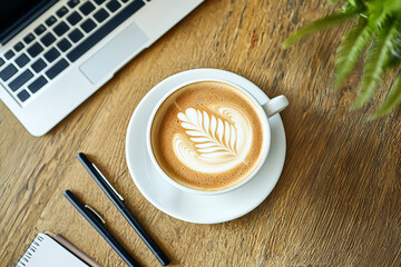 A modern workspace flat lay includes a laptop, coffee with latte art, smartphone, headphones, notebook, and pen on a wooden desk, illuminated by soft natural lighting