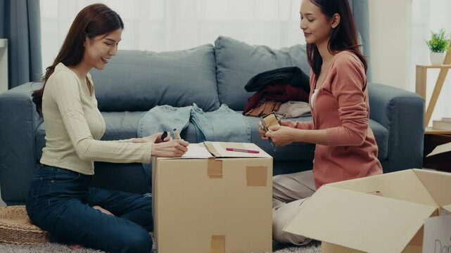 Two Asian women sort and pack clothes into donation boxes at home, contributing to charity and promoting sustainability through decluttering and reuse.